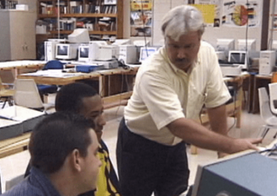 Male teacher in adult computer class room with two male students looking at a screen