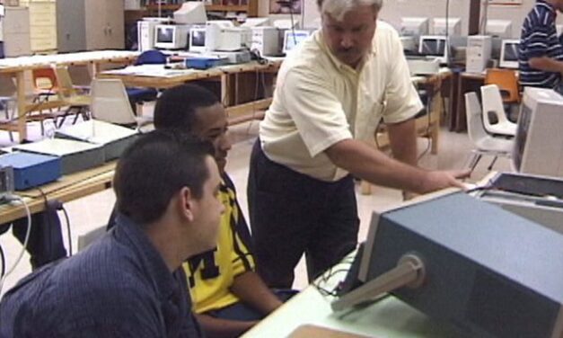 Male teacher in adult computer class room with two male students looking at a screen