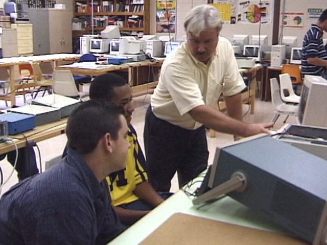Male teacher in adult computer class room with two male students looking at a screen