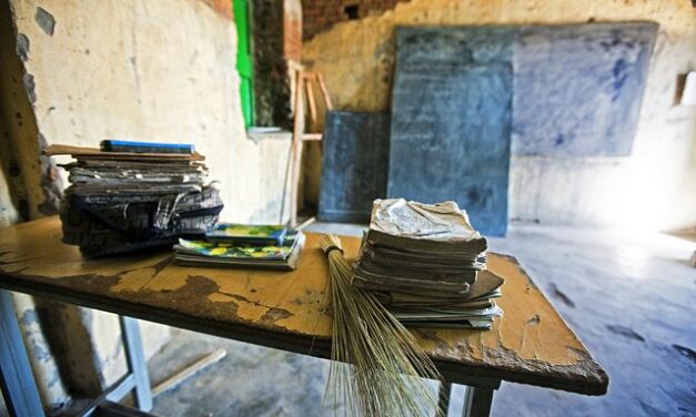 A teacher's desk in an empty classroom. Desk has books, and a cane on it