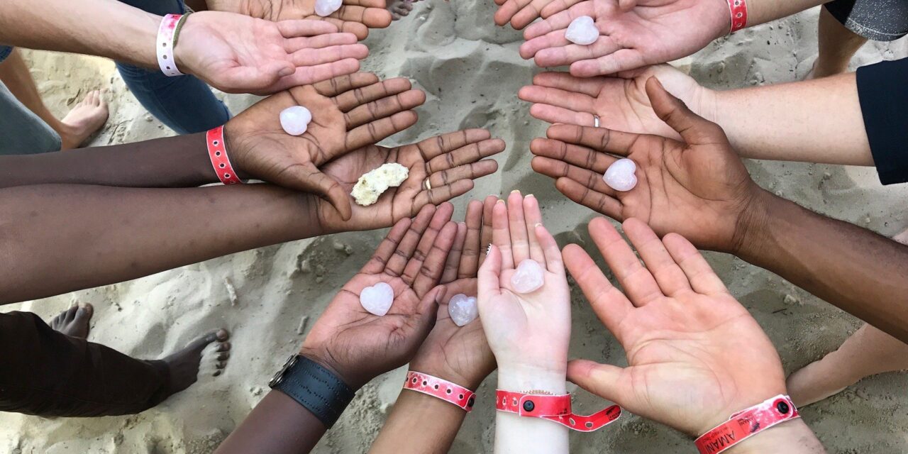 A ring of hands all with palms upturned, different coloured skin, some with a heart shape in the palm, some with nothing, one with coral