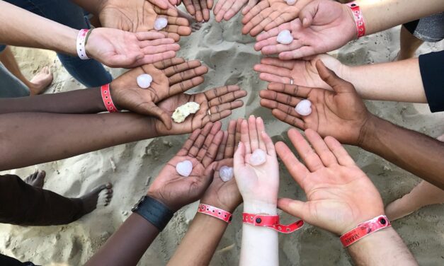 A ring of hands all with palms upturned, different coloured skin, some with a heart shape in the palm, some with nothing, one with coral