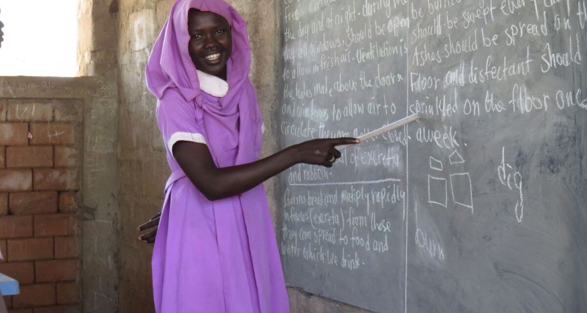 A woman in a pink dress at a blackboard at a camp in South Sudan