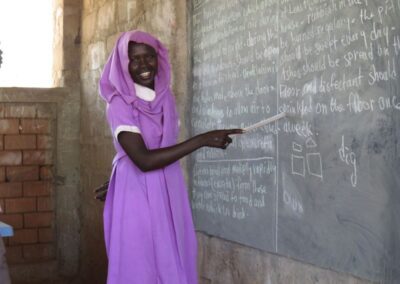 A woman in a pink dress at a blackboard at a camp in South Sudan