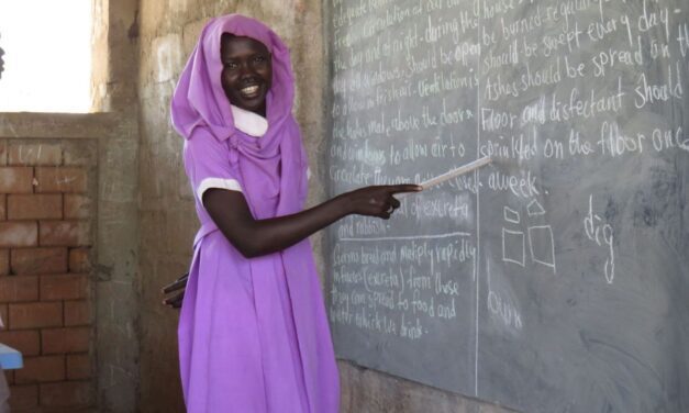 A woman in a pink dress at a blackboard at a camp in South Sudan