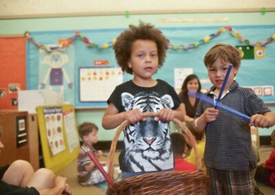 Boy with a basket in a primary school classroom, another boy has blue sticks