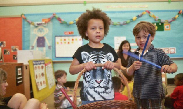 Boy with a basket in a primary school classroom, another boy has blue sticks