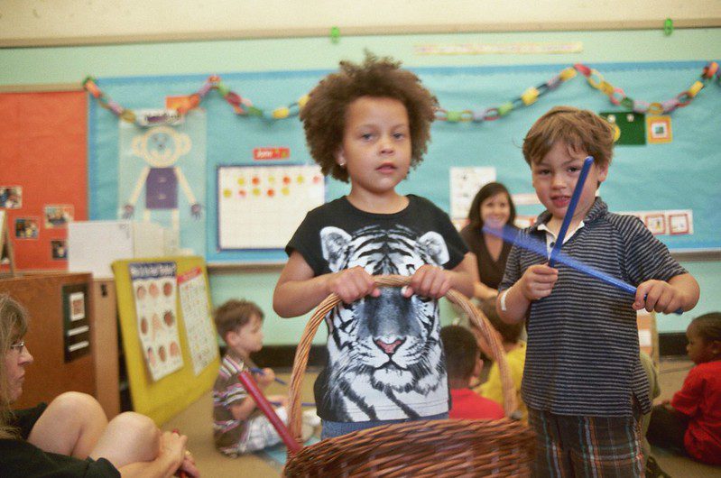 Boy with a basket in a primary school classroom, another boy has blue sticks