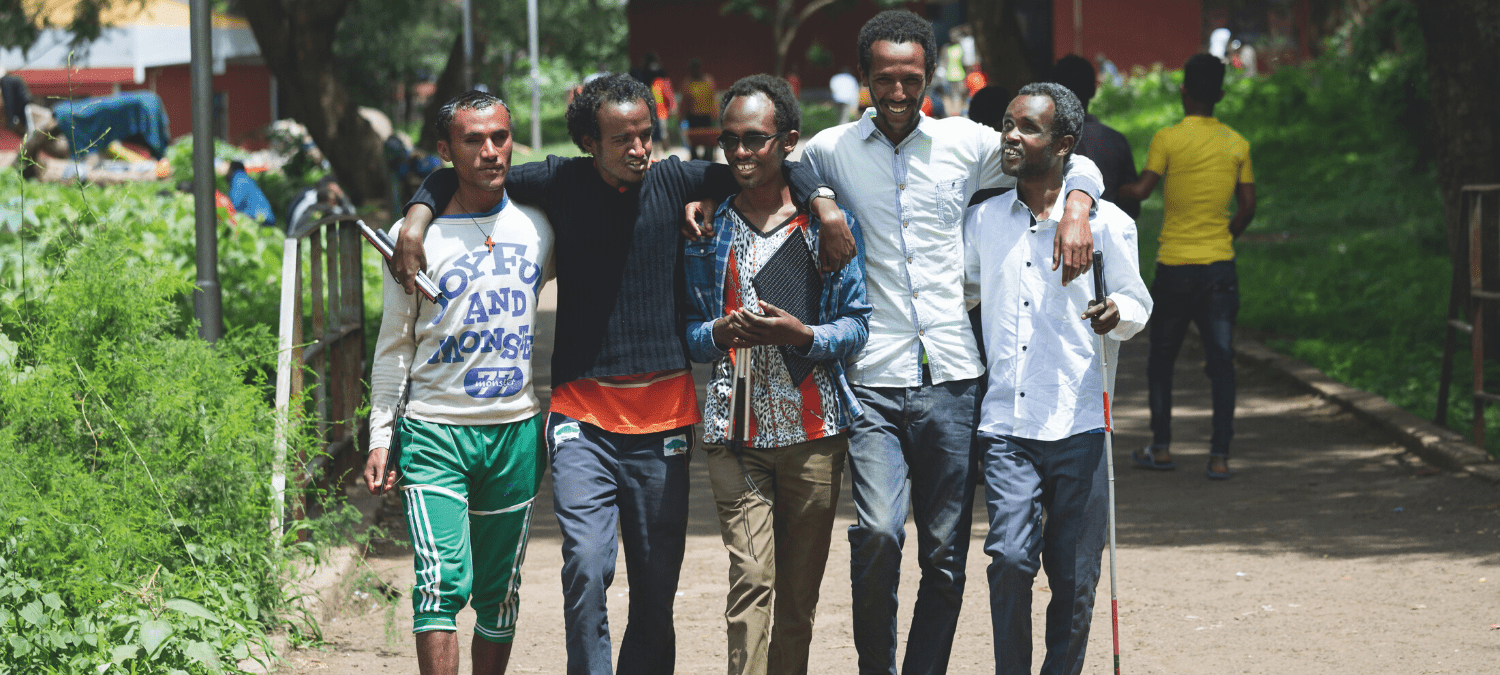 A group of five men walking towards the camera, linked together at their shoulders. Some are carrying canes to assist with visual imparement