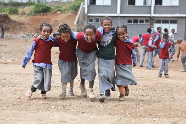 Five school children linking arms at the shoulders running in a row towards the camera. other school children in the background