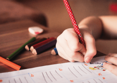 child's hand holding a pencil and marking on a page, coloured pencils scattered on the table