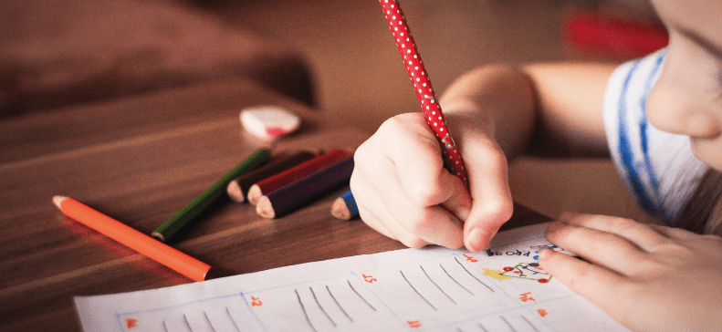 child's hand holding a pencil and marking on a page, coloured pencils scattered on the table