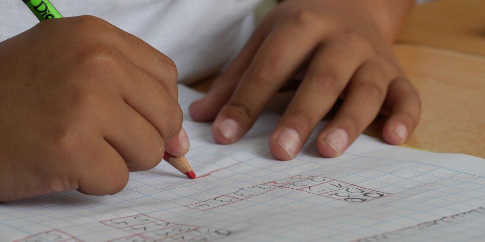 Child's hands holding a red coloured pencil working a number grid on squared paper
