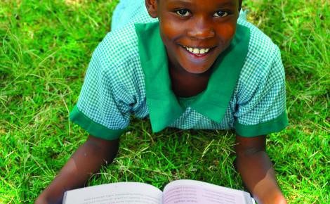 School girl in green uniform lying on her front on grass reading a book