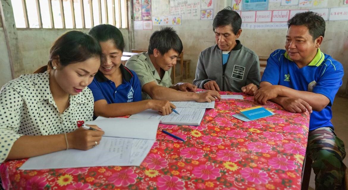 Teachers, the school principal and the village chief meet to discuss school finances and priorities. They are sitting around a table with a brightly coloured flowery cloth