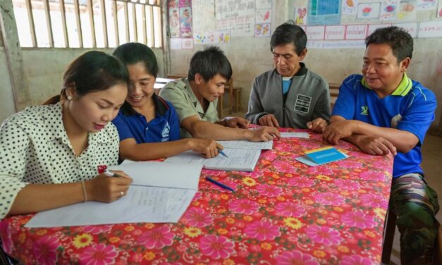 Teachers, the school principal and the village chief meet to discuss school finances and priorities. They are sitting around a table with a brightly coloured flowery cloth