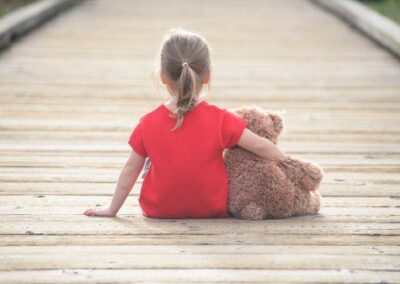 Young girl in red t shirt sitting alone on a boardwalk with her arm around a teddy bear