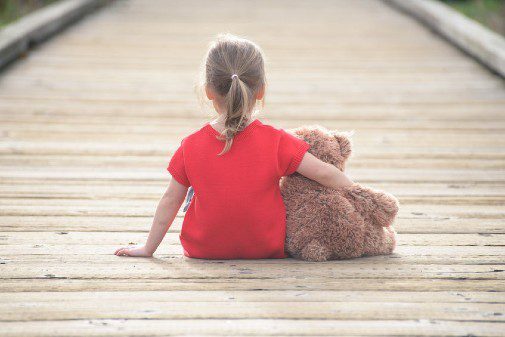 Young girl in red t shirt sitting alone on a boardwalk with her arm around a teddy bear