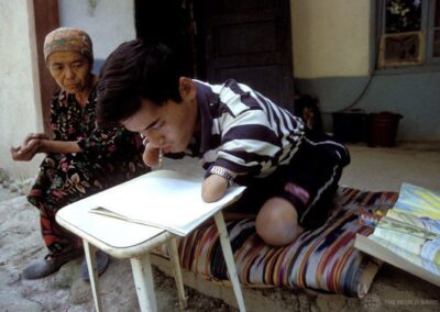 A boy with no forearms and no legs below the knew working with a pencil and paper outside a house. An older woman is with him