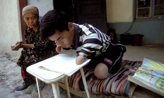 A boy with no forearms and no legs below the knew working with a pencil and paper outside a house. An older woman is with him