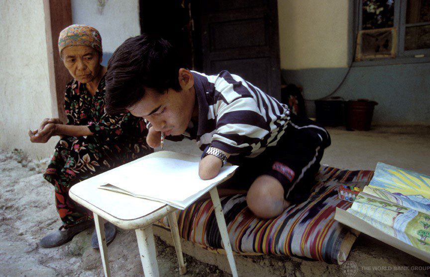 A boy with no forearms and no legs below the knew working with a pencil and paper outside a house. An older woman is with him