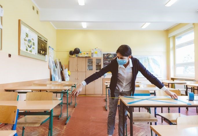 Teacher wearing a face mask measuring space between desks and marking some as do not sit here to make it covid safe