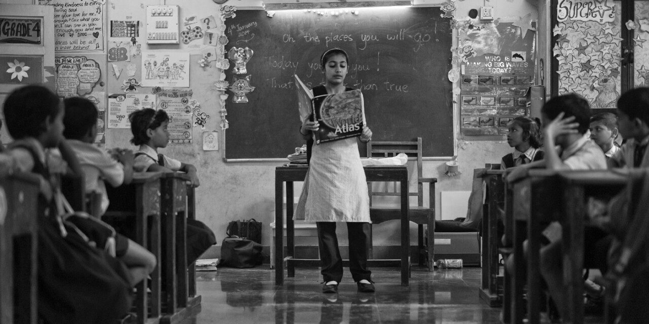 Black and white photo of teacher at front of classroom with an atlas