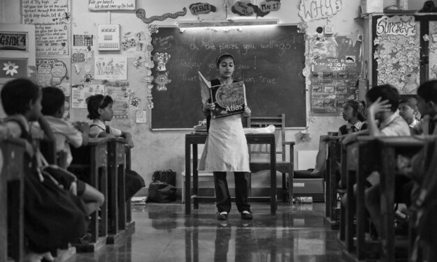 Black and white photo of teacher at front of classroom with an atlas