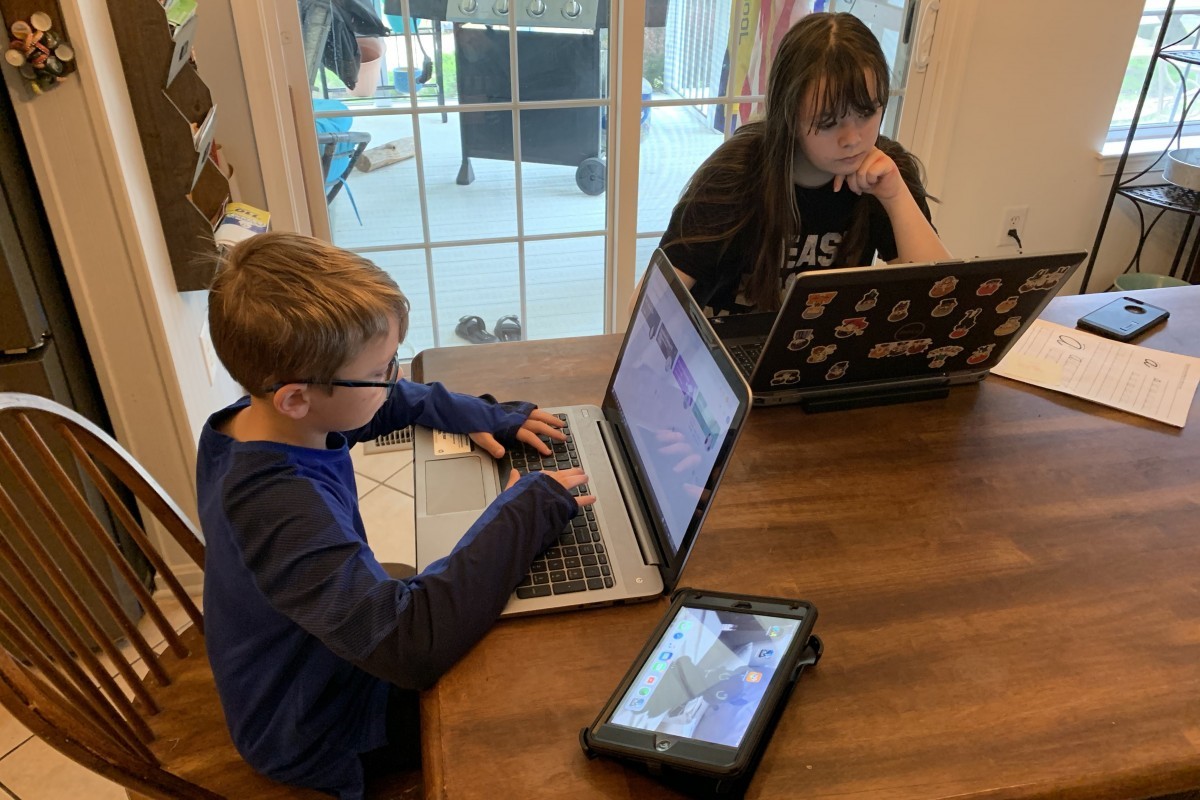 A boy and a girl with laptops at their dining room table also a tablet device on the table