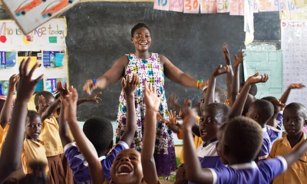 Teacher in primary classroom, smiling with her arms outstretched, the children are waving their hands in the air and shouting