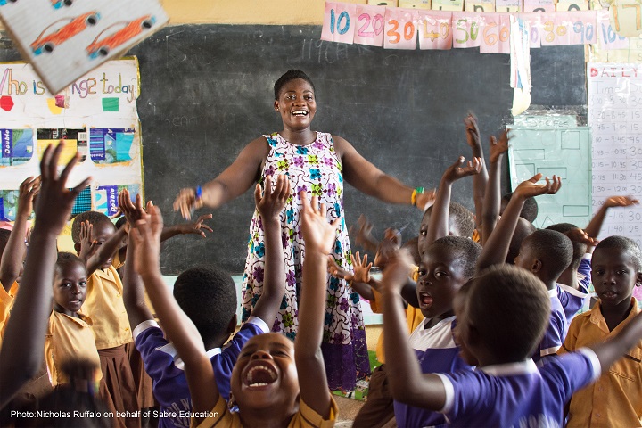 Teacher in primary classroom, smiling with her arms outstretched, the children are waving their hands in the air and shouting