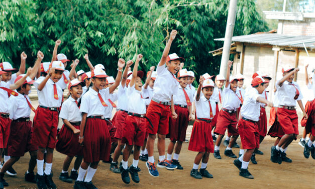 Asian children in school uniform - white shirt and dark red skirt/shorts jumping with hands raised outside a school