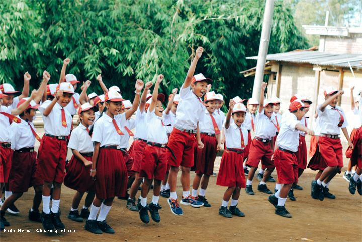 Asian children in school uniform - white shirt and dark red skirt/shorts jumping with hands raised outside a school