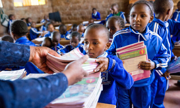 Primary age children in class wearing blue tracksuits as uniform. Three children are collecting stacks of text books from the teacher