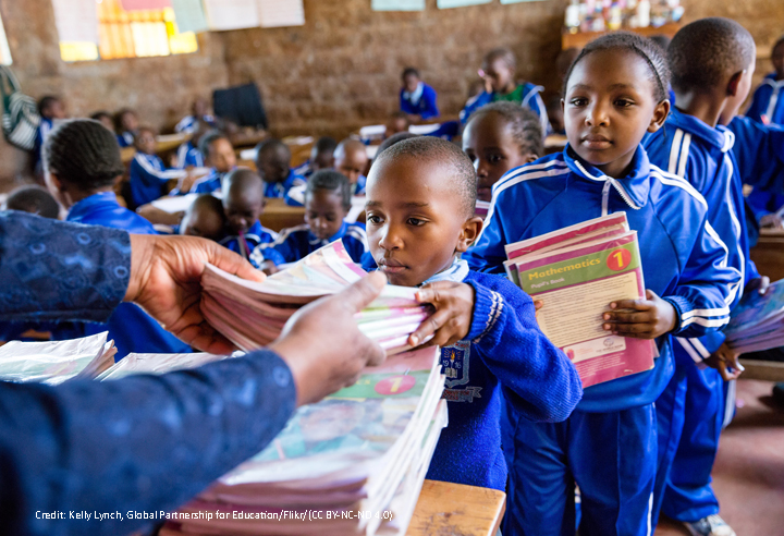Primary age children in class wearing blue tracksuits as uniform. Three children are collecting stacks of text books from the teacher