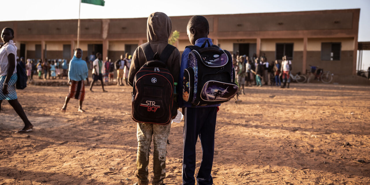 Two boys standing in the yard area of an African school, both wearing back packs. In the background is the school with other children assembling