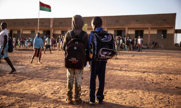 Two boys standing in the yard area of an African school, both wearing back packs. In the background is the school with other children assembling