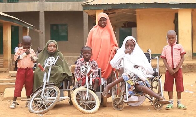 A group of women and children, some in wheelchairs or mobility bikes, outside a school. Two of the children have hearing aids