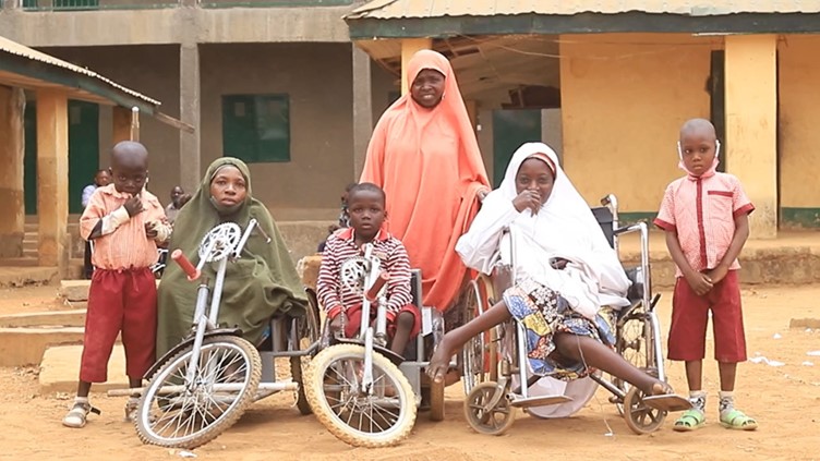 A group of women and children, some in wheelchairs or mobility bikes, outside a school. Two of the children have hearing aids