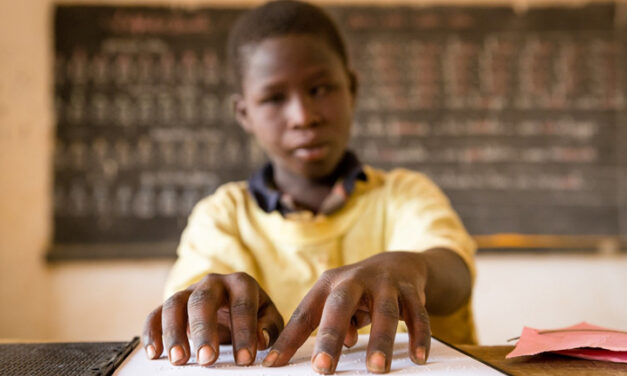 Blind child in a yellow top reading braille in a classroom