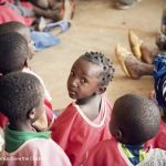 A group of African children sitting on the ground with feet of adults in view