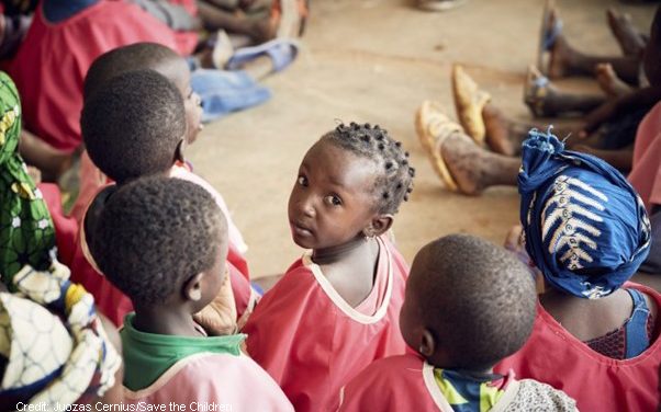 A group of African children sitting on the ground with feet of adults in view