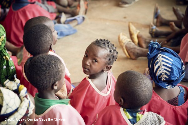 A group of African children sitting on the ground with feet of adults in view