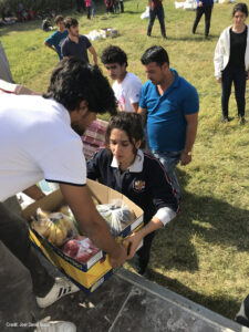 Students collaborate to deliver donations of food to a local refugee camp, Iraq. A girl takes a box of food from a person distributing, others wait behind