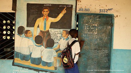 A student looking into his classroom, Sierra Leone, January 2015. On the wall by the door is a mural of a teacher and class with a child with a speech bubble - xxx is a very good maths teacher he makes us enjoy his lessons