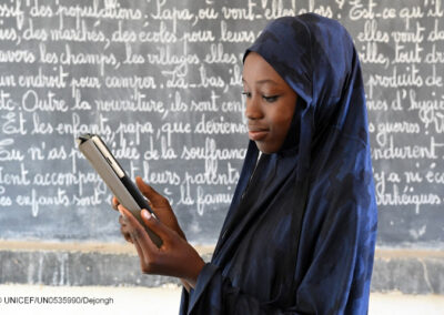 African girl with headdress looks at a tablet device in front of a blackboard in a classroom.
