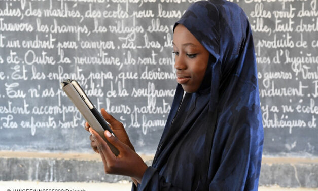 African girl with headdress looks at a tablet device in front of a blackboard in a classroom.