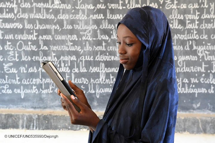 African girl with headdress looks at a tablet device in front of a blackboard in a classroom.