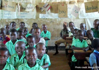 Primary school children dressed in green school uniforms sit in an over-crowded classroom in Uganda, with sheets of paper hanging across the ceiling of English words they are learning to write.