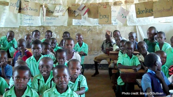 Primary school children dressed in green school uniforms sit in an over-crowded classroom in Uganda, with sheets of paper hanging across the ceiling of English words they are learning to write.
